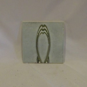 May include: A light blue ceramic tile with a green stylized design of an oval shape with a series of curved lines.