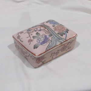 May include: A small, rectangular porcelain trinket box with a pink base and a floral design. The box features a blue and white bird with pink, blue, and white flowers.