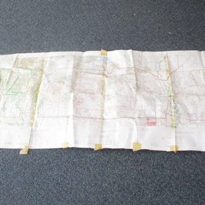 May include: A folded map, secured with tape at the corners, lies on a dark gray surface. The map is white with various colored lines and markings, suggesting a road map or topographical chart. The map is creased.