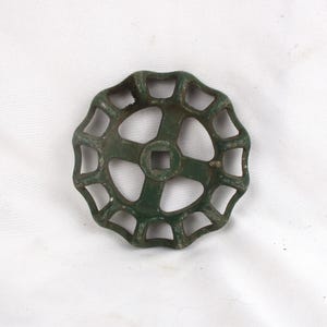 May include: A vintage, circular valve handle with a dark green, weathered finish. The handle features an open, spoke-like design with a square center hole. The metal shows signs of age and use, suggesting it is an antique.