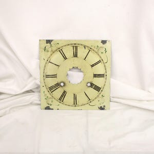 May include: Square clock face with Roman numerals and a central hole. The face is off-white with black numerals and minute markers. Decorative green floral accents are in each corner.