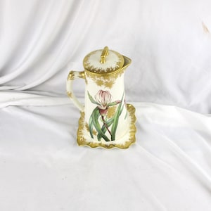 May include: A vintage porcelain coffee pot with a lid, handle, and a matching base. The pot is white with gold trim and features a floral design with pink and green accents. The pot is approximately 8 inches tall.