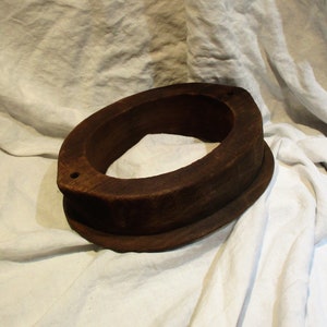 May include: A dark brown wooden hat form with a rounded oval shape and two small holes on the side. The form is sitting on a white fabric surface.