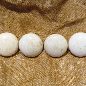 May include: Four off-white, round cabinet knobs with slight variations in color and texture. The knobs are arranged in a row against a textured, tan burlap background. These knobs could be used for drawers or cabinets.