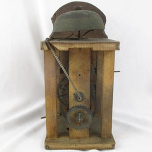 May include: A vintage wooden clock mechanism with visible gears and a bell. The clock's wooden frame has a natural finish, and the gears are made of metal. The bell is a dark green color and is mounted on top of the clock.