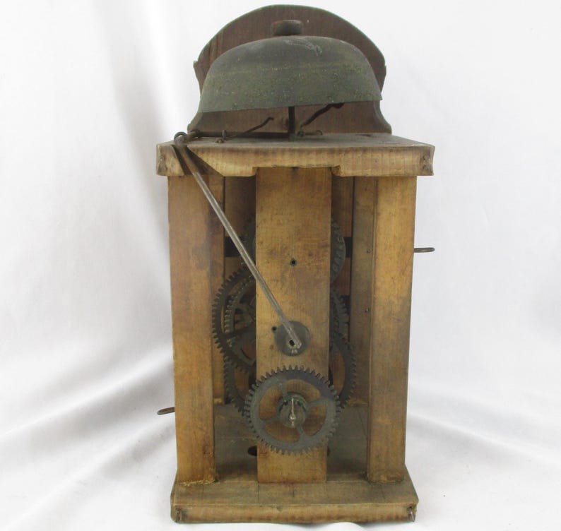 May include: A vintage wooden clock mechanism with visible gears and a bell. The clock's wooden frame has a natural finish, and the gears are made of metal. The bell is a dark green color and is mounted on top of the clock.