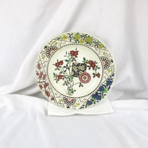 May include: A decorative plate with a floral design. The plate features a cream-colored center with red, yellow, and blue floral patterns around the rim. The plate has a diameter of approximately 9 inches.