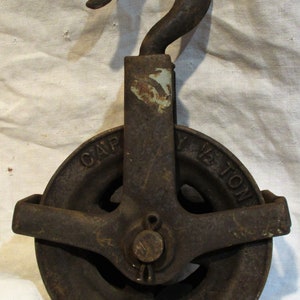Pu&ograve; includere: Una carrucola in ghisa vintage con un gancio, contrassegnata "CAPY 1/2 TON". La carrucola &egrave; arrugginita e presenta una patina marrone scuro.