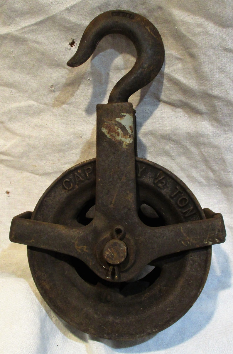 Pu&ograve; includere: Una carrucola in ghisa vintage con un gancio, contrassegnata "CAPY 1/2 TON". La carrucola &egrave; arrugginita e presenta una patina marrone scuro.