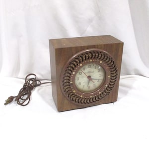 May include: A vintage electric clock with a square wooden frame and a circular clock face. The clock face has a light-colored background with black numerals and hands. The clock is brown and has a decorative, circular border around the clock face. A brown power cord extends from the clock.