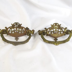 May include: Two antique brass drawer pulls with ornate, filigree designs. The pulls have a curved handle and a decorative backplate. The brass has a warm, aged patina, suggesting vintage or antique origins. These pulls would be suitable for furniture restoration.