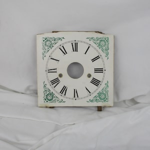 May include: White clock face with black Roman numeral markings and green floral accents. The clock face is square and has a hole in the center for the clock hands.