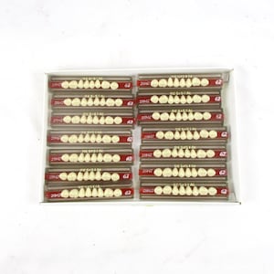 May include: A rectangular box containing multiple rows of artificial teeth. Each row is in a small, labeled container with the text "RESISTAL" and the number "62". The teeth are a creamy white color.