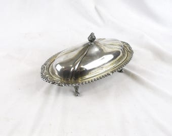 May include: A silver-colored, oval-shaped butter dish with a lid and ornate detailing. The dish has a decorative edge and sits on small, curved feet. The lid features a small, textured handle. The butter dish is likely made of metal.