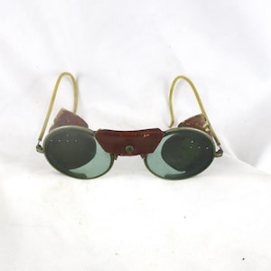 May include: Vintage goggles with round, green-tinted lenses and a brown leather bridge. The frames are a brass color, and the ear pieces are a light yellow. The goggles are set against a white background.