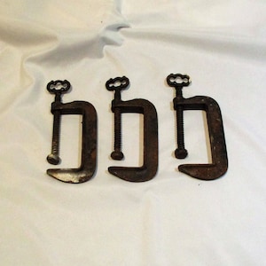 May include: Three rusty metal C-clamps with a screw adjustment. The clamps are arranged in a row, with the handles facing the viewer.