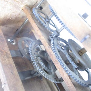 May include: Close-up of the inner workings of a wooden clock. The image shows a series of metal gears, wheels, and a circular disc, all set within a wooden frame. The gears are intricately designed and appear to be in good condition.