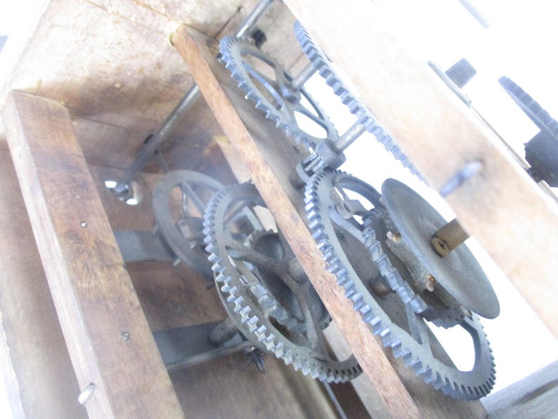 May include: Close-up of the inner workings of a wooden clock. The image shows a series of metal gears, wheels, and a circular disc, all set within a wooden frame. The gears are intricately designed and appear to be in good condition.
