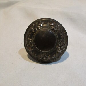 May include: A round, ornate, metal door knob with a decorative design. The knob is a dark brown color and has a smooth, circular center.