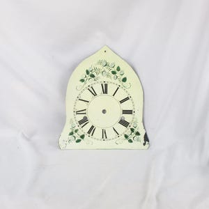 May include: Off-white clock face with black Roman numerals and hands. The clock face has a decorative floral design in green and white around the perimeter. The clock face is an arched shape with a small hole at the top.