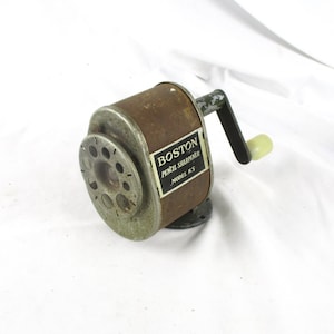 May include: Vintage Boston Pencil Sharpener Model KS. The sharpener has a cylindrical body with a crank handle and a dial for pencil insertion. The body is a mix of brown and green with the text "BOSTON PENCIL SHARPENER MODEL KS" on a label.