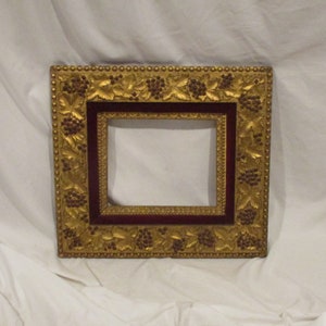 May include: A gold picture frame with a burgundy velvet mat and a grapevine design. The frame is ornate and has a beaded edge.
