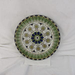 May include: A white ceramic plate with a blue and green floral design. The plate has a decorative border with a repeating pattern of green leaves and gold accents.