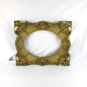 May include: Ornate, gold-colored oval picture frame with detailed scrollwork and decorative flourishes. The frame has a vintage aesthetic and is designed to hold an oval-shaped image. The frame is made of a gold-colored material.