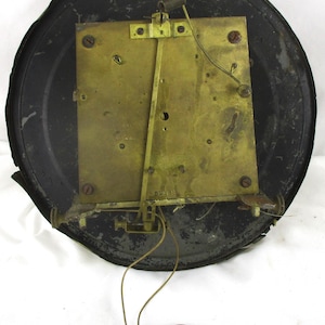 Peut inclure: Le dos d'un m&eacute;canisme d'horloge vintage. Le cadre m&eacute;tallique de l'horloge est noir et circulaire, avec une plaque rectangulaire de couleur laiton au centre. Un pendule et des fils sont suspendus au m&eacute;canisme. Le cadre de l'horloge pr&eacute;sente des signes d'usure.
