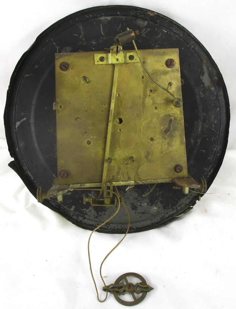 Peut inclure: Le dos d'un m&eacute;canisme d'horloge vintage. Le cadre m&eacute;tallique de l'horloge est noir et circulaire, avec une plaque rectangulaire de couleur laiton au centre. Un pendule et des fils sont suspendus au m&eacute;canisme. Le cadre de l'horloge pr&eacute;sente des signes d'usure.