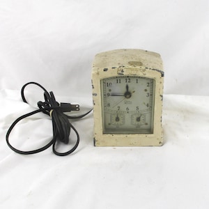May include: An antique cream-colored electric clock with a rectangular face and a curved top. The clock face has black numerals and hands, with two smaller dials below. A black power cord is attached.