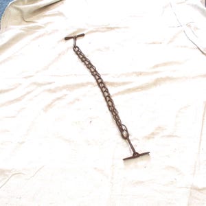 May include: A rusty brown metal chain with two T-shaped ends. The chain is made of small, interconnected links. The object is lying on a white, textured surface.