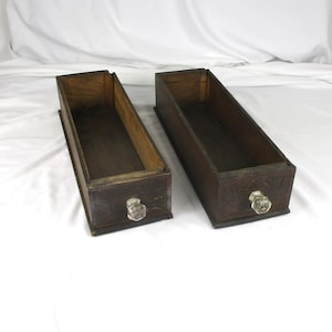 May include: Two rectangular wooden drawers with glass knobs. The drawers are dark brown with a lighter brown interior. The drawers are empty and sit side by side.