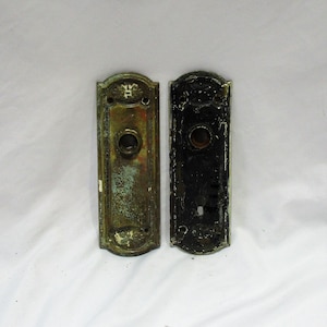 May include: Two antique door plates, one brass and one black, with a keyhole and a round knob. Both plates have decorative details.