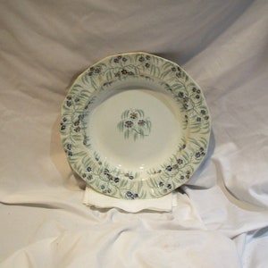 May include: A white ceramic soup bowl with a green and blue floral pattern around the rim. The bowl has a scalloped edge and a floral design in the center.