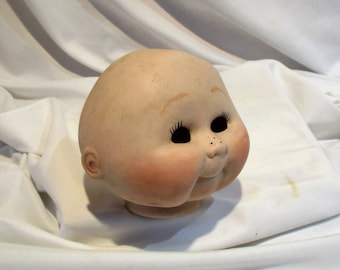 May include: A porcelain doll head with painted features, including large eyes, rosy cheeks, and a small nose. The head is designed for doll making and has a blank expression.