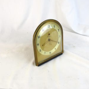 May include: A vintage brass-toned clock with a curved top and a rectangular base. The clock face is a light gold color with black numerals and hands. The clock is set against a white background.