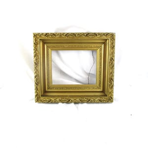 May include: A rectangular picture frame with a metallic gold finish. The frame features an ornate, raised relief pattern along the outer border and a smooth, recessed inner section. A thin, dark wire is visible inside the open center area.