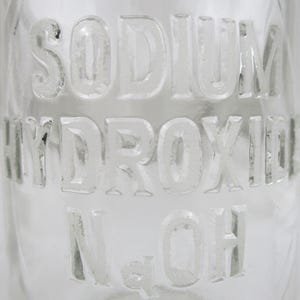 Może przedstawiać: Przezroczysta szklana butelka z wytłoczonymi słowami "SODIUM HYDROXIDE" i "NaOH". Litery są wypukłe i w stylu bezszeryfowym. Przejrzystość butelki pozwala na zobaczenie tła.