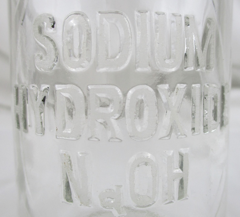 Może przedstawiać: Przezroczysta szklana butelka z wytłoczonymi słowami "SODIUM HYDROXIDE" i "NaOH". Litery są wypukłe i w stylu bezszeryfowym. Przejrzystość butelki pozwala na zobaczenie tła.