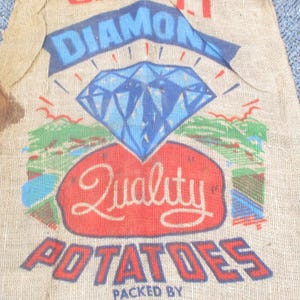 May include: A tan burlap sack featuring a blue diamond graphic above a red oval with the word Quality in white script. Below this, the word POTATOES is printed in large blue letters with red outlines, and the text PACKED BY is visible at the bottom.