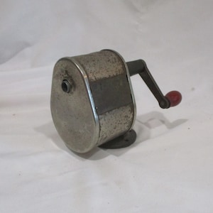 May include: A vintage metal pencil sharpener with a crank handle and a red knob. The sharpener has a textured surface and a silver finish.