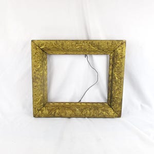 May include: A rectangular, antique gold picture frame with ornate floral detailing. The frame is empty, revealing a white background. A thin, black wire is visible within the frame. The frame's design suggests a vintage or classic aesthetic.