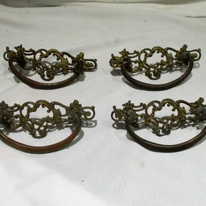 May include: Four antique brass drawer pulls with ornate, scrolled designs. The pulls are in the shape of a half-circle with a decorative loop at the top.