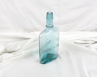 May include: A vintage, light blue glass bottle with a rounded body and a narrow neck. The bottle has embossed text that reads "GILBERT BROS & BALTIMORE MD". The bottle is empty and sits on a white surface.