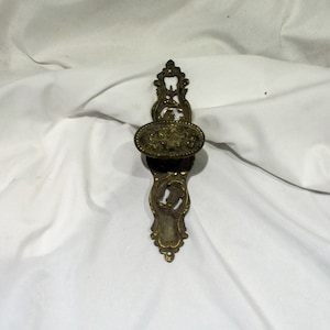 May include: A vintage brass door handle with an ornate design. The handle is oval shaped and has a decorative floral pattern. The handle is attached to a long, rectangular plate with a similar floral design.