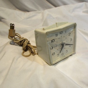 May include: A white electric alarm clock with a black face and hands. The clock has a cord with a plug.