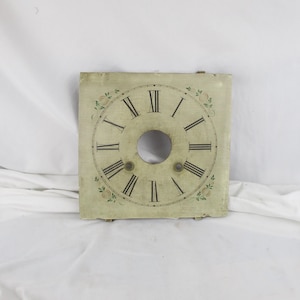 May include: Square clock face with Roman numerals and a central hole. The face is off-white with black numerals and hands, and decorative floral accents. The clock face is likely for a vintage or antique clock.