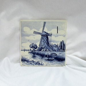 May include: Blue and white Delft tile featuring a windmill with a canal and trees in the background. The tile has a blue and white design with a white background.