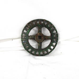 May include: A weathered, circular metal object with a central square hole and a cross-shaped spoke design. The outer rim features evenly spaced rectangular cutouts. The object has a distressed, aged appearance with a green and brown patina.